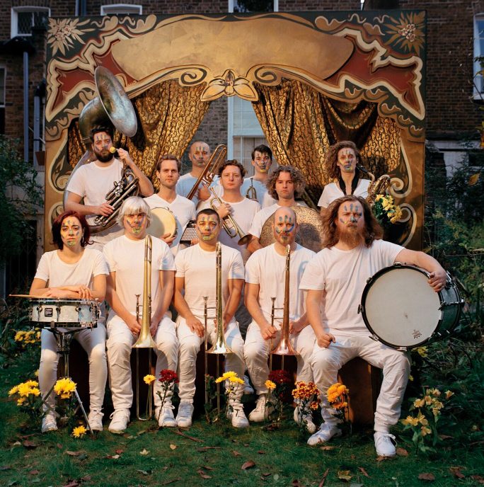 Hackney Colliery band - a group of 12 people all dressed in white facing the camera, sitting a differnt heights holding musical instruments