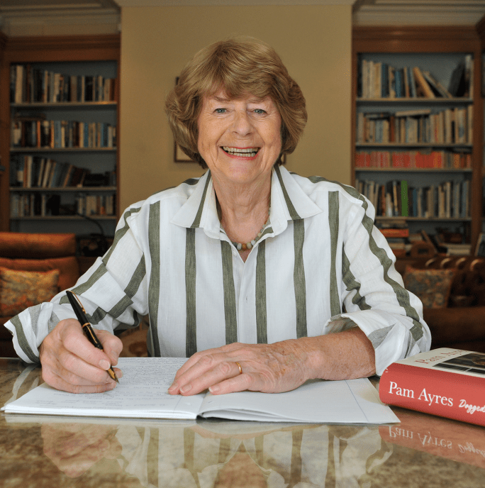 Poet Pam Ayres, an older lady sat a desk holding a pen writing in a book. she has short hair and is wearing a white and green stripped shirt
