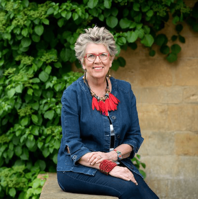 Dame Prue Leith DBE. Woman with short hair sitting on a wall wearing a denim jacket, a bold birght red necklace and red glasses