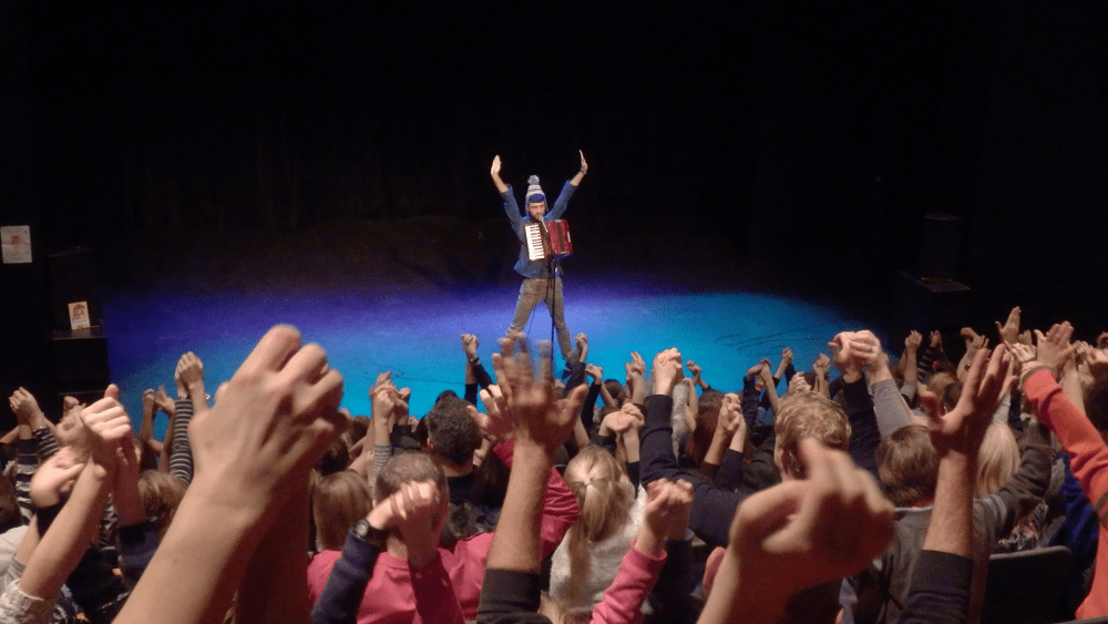 comedy club 4 kids. a performer on stage with a his hands in the air. Photo is take from the back of the room, the audience all have their hands in the air