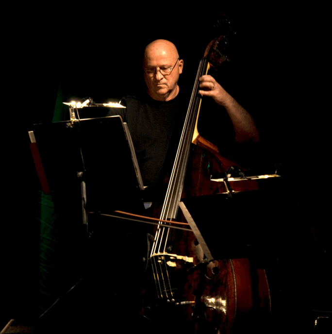 Gavin Bryars sits in a chair holding a large musical instrument he has a music stand in front of him. He is wearing all black and is against a black background