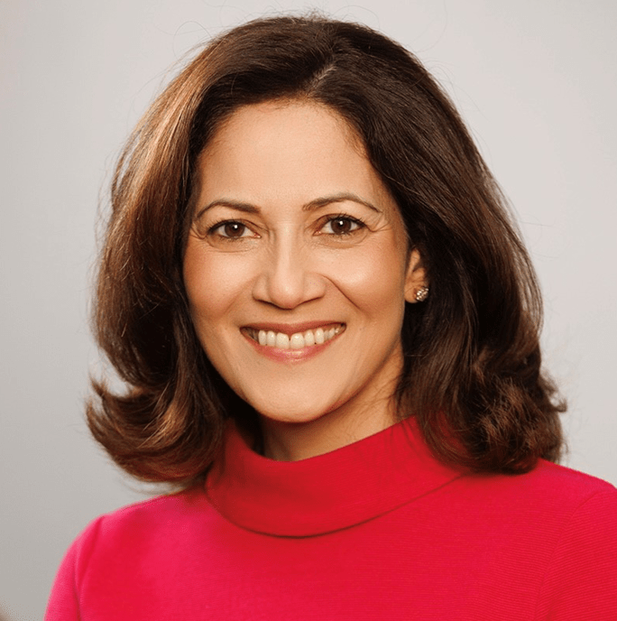 a headshot of Mishal Husain against a grey background. she is waering a red jumper and has short brown hair